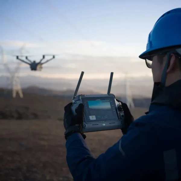 DJI Matrice 4D im Flug mit RC Plus im Vordergrund und Hochspannungsleitungen im Hintergrund